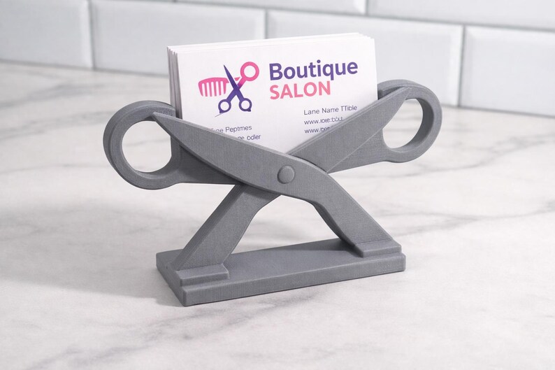 May include: A gray scissor-shaped business card holder. The holder has a base and two large scissor blades with circular handles. White business cards with the text "Boutique Salon" are in the holder.