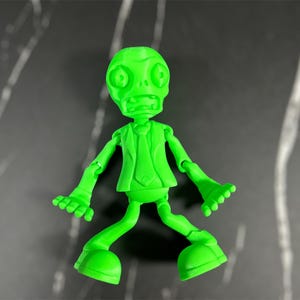 May include: A green plastic toy figure of a zombie wearing a suit and tie. The figure has a wide open mouth and large eyes. The figure is posed with its arms outstretched and its legs spread apart.