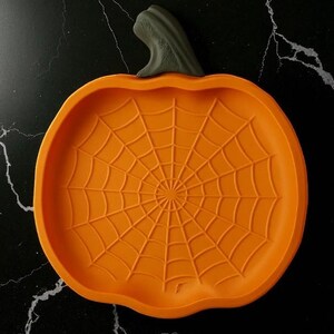 May include: Orange pumpkin-shaped serving dish with a spiderweb design. The dish has a dark gray stem and is set against a black background. A festive Halloween-themed item.