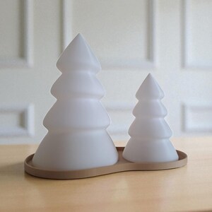 May include: Two frosted white Christmas tree-shaped decorative lights sit on a light brown wooden base. The larger tree is on the left, and the smaller tree is on the right. The trees have a layered design.