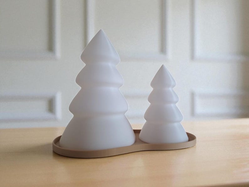 May include: Two frosted white Christmas tree-shaped decorative lights sit on a light brown wooden base. The larger tree is on the left, and the smaller tree is on the right. The trees have a layered design.