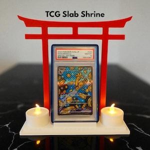 May include: A red TCG Slab Shrine displaying a Pokemon card in a protective case. The card features a colourful illustration of a Gholdengo ex Special Art Rare card. Two white candles flank the card, and the text "TCG Slab Shrine" is at the top.