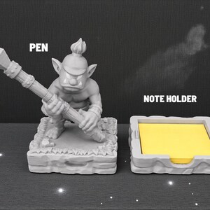 May include: A gray pen holder shaped like a fantasy creature holding a spear, with the word "PEN" above it. Next to it is a gray note holder with yellow sticky notes and the words "NOTE HOLDER".