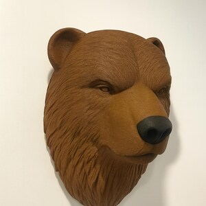 Puede incluir: Escultura de cabeza de oso marr&oacute;n con nariz negra y textura de pelaje detallada. La cabeza del oso est&aacute; montada en una pared blanca, mostrando sus caracter&iacute;sticas realistas y artesan&iacute;a. Es un art&iacute;culo decorativo.