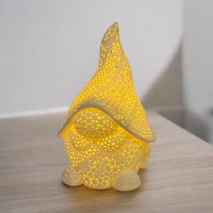 May include: A ceramic gnome-shaped lamp with a yellow glow. The lamp has a pointed hat and a round body, both covered in small, evenly spaced holes. The light shines through the holes, creating a warm, inviting ambiance. The lamp is sitting on a wooden surface.