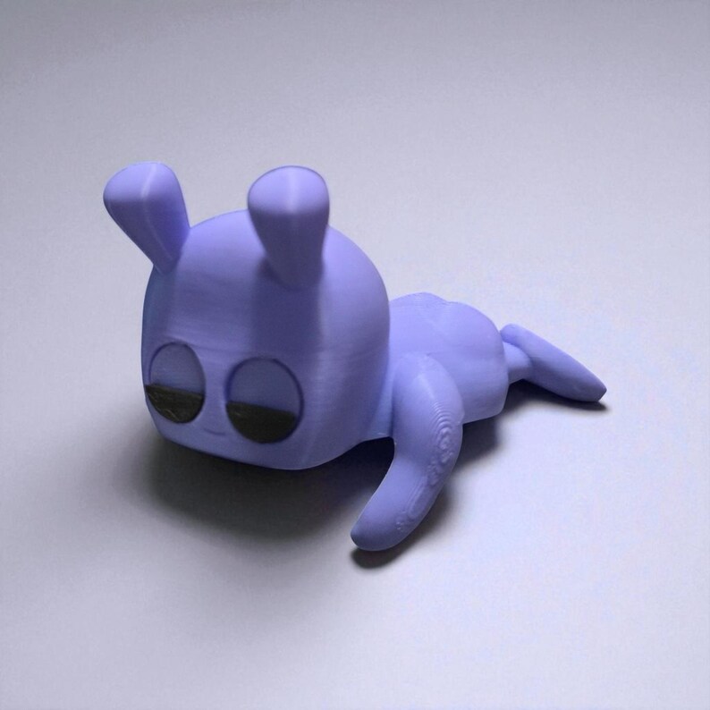 Pu&ograve; includere: Una statuetta lavanda stampata in 3D di una creatura stilizzata con una testa squadrata, grandi occhi e piccole orecchie. La creatura &egrave; sdraiata con le braccia e le gambe distese. Gli occhi sono neri.