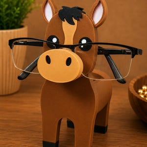 May include: A brown horse-shaped eyeglasses holder with black-framed glasses. The holder has a light brown muzzle and ears, black eyes, and a black mane. The holder is made of wood and sits on a wooden surface.