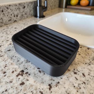 May include: A rectangular black soap dish with a slatted top. The dish is made of a matte black material and is designed to hold a bar of soap. The dish measures approximately 5 inches long and 3 inches wide.