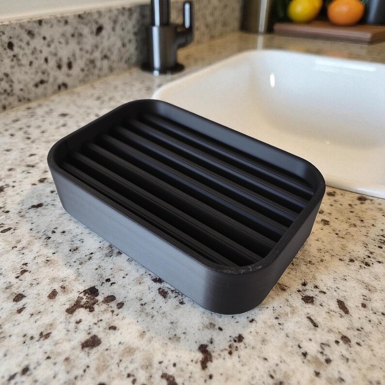 May include: A rectangular black soap dish with a slatted top. The dish is made of a matte black material and is designed to hold a bar of soap. The dish measures approximately 5 inches long and 3 inches wide.