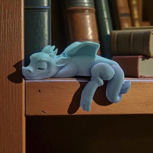 May include: A light blue dragon figurine resting on a wooden shelf. The dragon has closed eyes, wings, and detailed scales. The background features a collection of books, suggesting a fantasy or literary theme.
