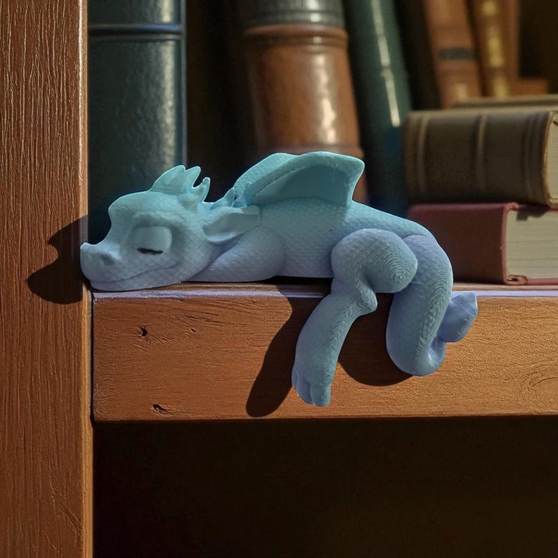 May include: A light blue dragon figurine resting on a wooden shelf. The dragon has closed eyes, wings, and detailed scales. The background features a collection of books, suggesting a fantasy or literary theme.