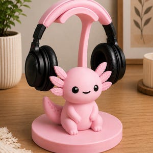 May include: A pink axolotl-shaped headphone stand with a pair of black headphones resting on top. The stand is made of a solid pink material and features a cute axolotl figurine at the base.