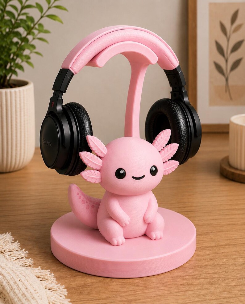May include: A pink axolotl-shaped headphone stand with a pair of black headphones resting on top. The stand is made of a solid pink material and features a cute axolotl figurine at the base.