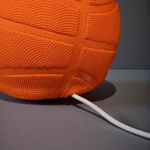 May include: Close-up of an orange basketball-shaped object with a textured surface and dark orange lines. A white cable extends from the object. The object is on a grey surface.