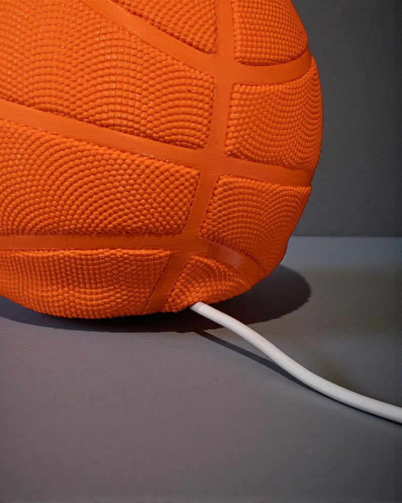 May include: Close-up of an orange basketball-shaped object with a textured surface and dark orange lines. A white cable extends from the object. The object is on a grey surface.