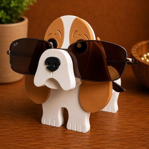 May include: A wooden dog-shaped sunglasses holder in white, brown, and black. The dog is wearing brown-tinted sunglasses. The holder is on a wooden surface.
