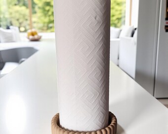 May include: A paper towel holder with a roll of white paper towels. The holder has a beige, scalloped base and a wooden knob on top. The paper towels have a geometric pattern. The holder is on a white countertop.