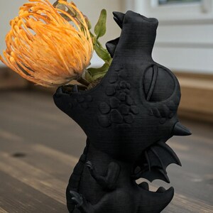 May include: A black dragon-shaped vase holding an orange protea flower. The vase has detailed scales, wings, and a textured finish. The flower's vibrant color contrasts with the dark vase, creating a striking decorative piece.