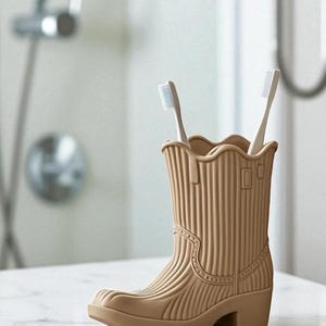 May include: A tan-colored, cowboy boot-shaped toothbrush holder with two white toothbrushes. The holder has a ridged texture and decorative stitching details. The boot is made of a durable material and is designed to hold toothbrushes upright in a bathroom setting.