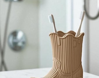 May include: A tan-colored, cowboy boot-shaped toothbrush holder with two white toothbrushes. The holder has a ridged texture and decorative stitching details. The boot is made of a durable material and is designed to hold toothbrushes upright in a bathroom setting.
