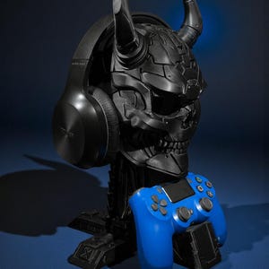 以下が含まれることがあります： 角のある頭蓋骨の形をした黒いゲーミングヘッドセットスタンド。ベースには青いゲームコントローラーが置かれています。スタンドは黒いワイヤレスヘッドホンを保持しています。デザインは未来的で機械的な美学を持っています。