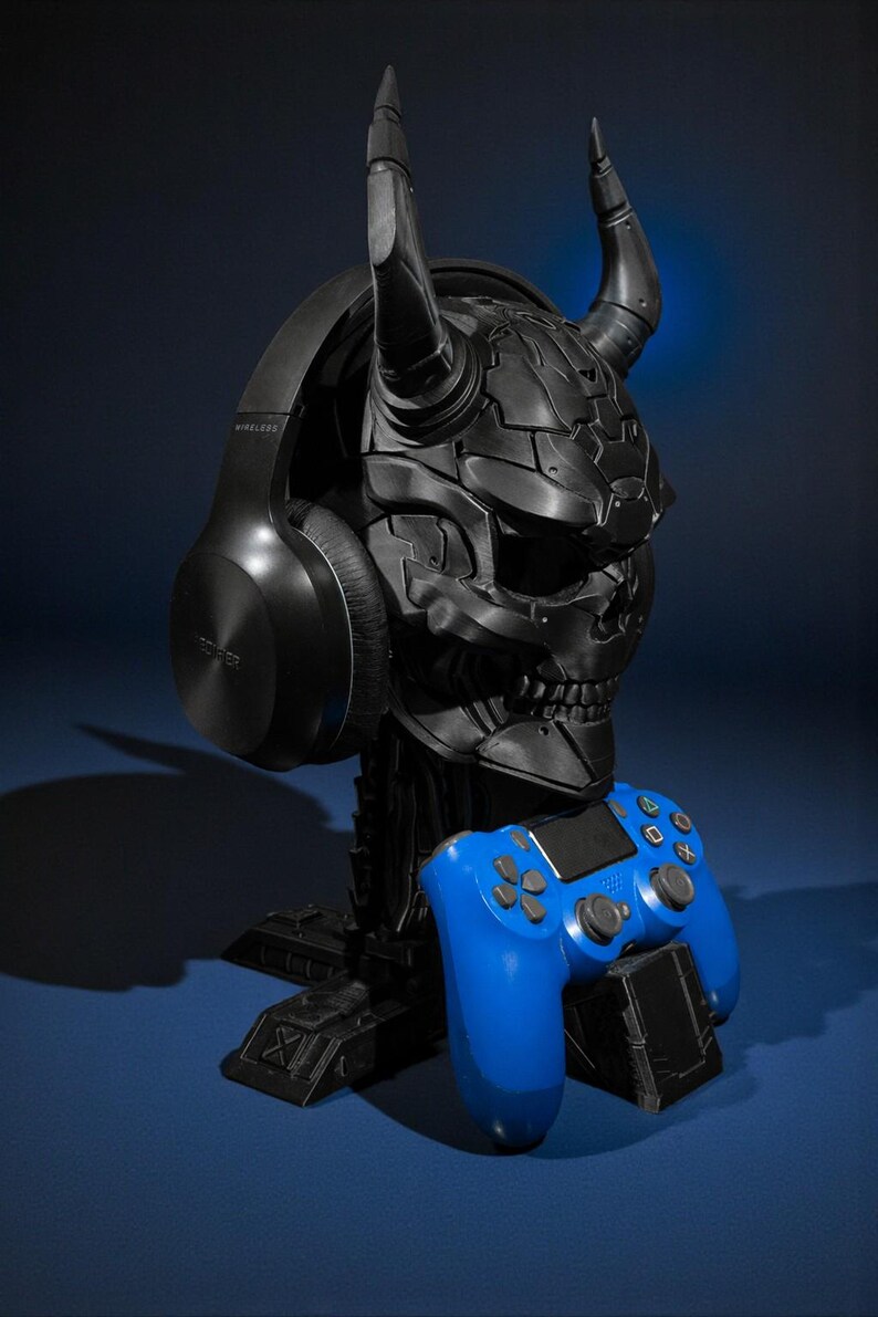 以下が含まれることがあります： 角のある頭蓋骨の形をした黒いゲーミングヘッドセットスタンド。ベースには青いゲームコントローラーが置かれています。スタンドは黒いワイヤレスヘッドホンを保持しています。デザインは未来的で機械的な美学を持っています。