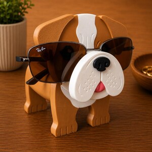 May include: A brown and white bulldog-shaped sunglasses holder with sunglasses. The holder has a white snout, black nose, and pink tongue. The sunglasses are dark-tinted with a silver frame. The holder is on a wooden surface.