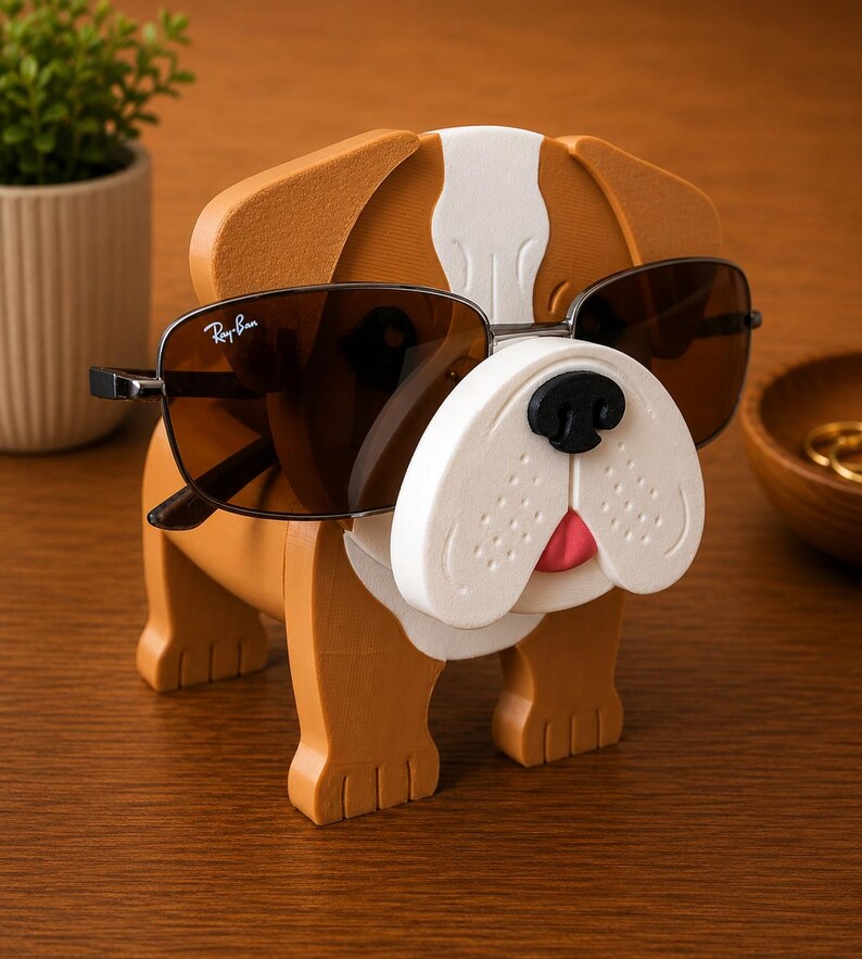 May include: A brown and white bulldog-shaped sunglasses holder with sunglasses. The holder has a white snout, black nose, and pink tongue. The sunglasses are dark-tinted with a silver frame. The holder is on a wooden surface.