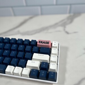 May include: A blue and white mechanical keyboard with a pink keycap that says "ERASE" in black lettering.