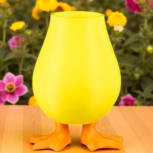 Puede incluir: Un jarr&oacute;n decorativo amarillo brillante con forma de pato. El jarr&oacute;n tiene un cuerpo redondeado y patas de pato naranjas como base. El fondo presenta una variedad de flores coloridas.