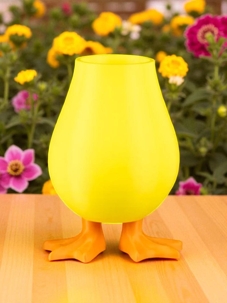 Puede incluir: Un jarr&oacute;n decorativo amarillo brillante con forma de pato. El jarr&oacute;n tiene un cuerpo redondeado y patas de pato naranjas como base. El fondo presenta una variedad de flores coloridas.