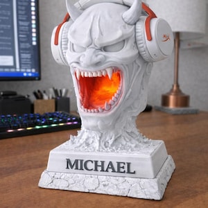 May include: A white, devil-themed desk lamp with a snarling face, horns, and headphones. The mouth glows orange. The base reads "MICHAEL". The headphones are white with red accents. A computer monitor and keyboard are in the background.