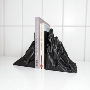 May include: Black mountain-shaped bookends holding two books. The bookends have a textured surface and are made of a solid material. The books are titled "THE STRENGTH IN OUR SCARS" and "KATHERINE MAY OBERWINTERN".