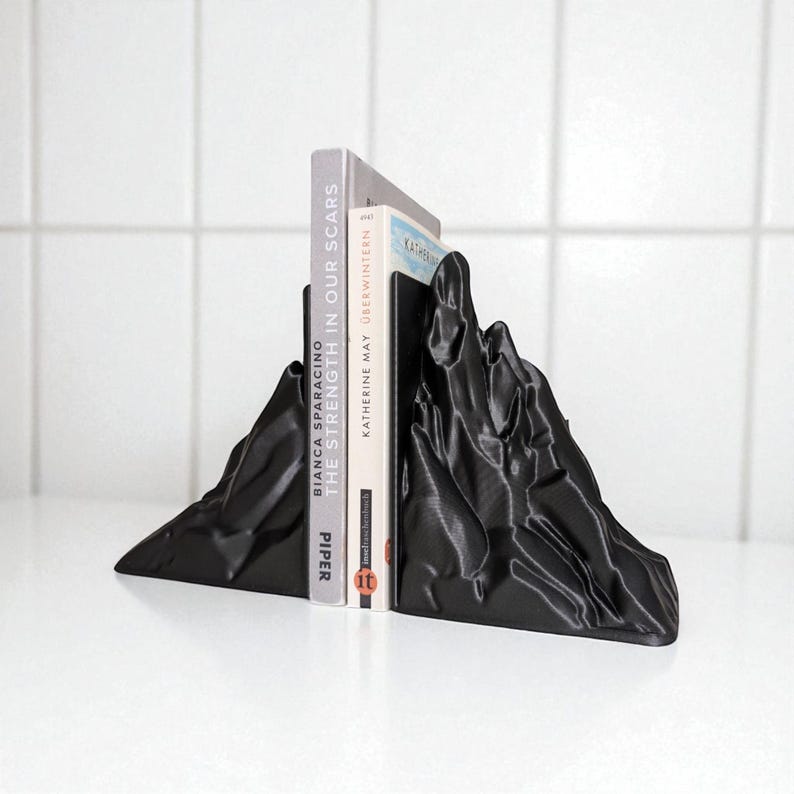 May include: Black mountain-shaped bookends holding two books. The bookends have a textured surface and are made of a solid material. The books are titled "THE STRENGTH IN OUR SCARS" and "KATHERINE MAY OBERWINTERN".