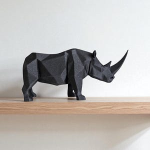 May include: A black, geometric rhino sculpture with a textured surface. The rhino is positioned on a light-colored wooden shelf against a white wall. The sculpture has a prominent horn and is a modern decorative piece.
