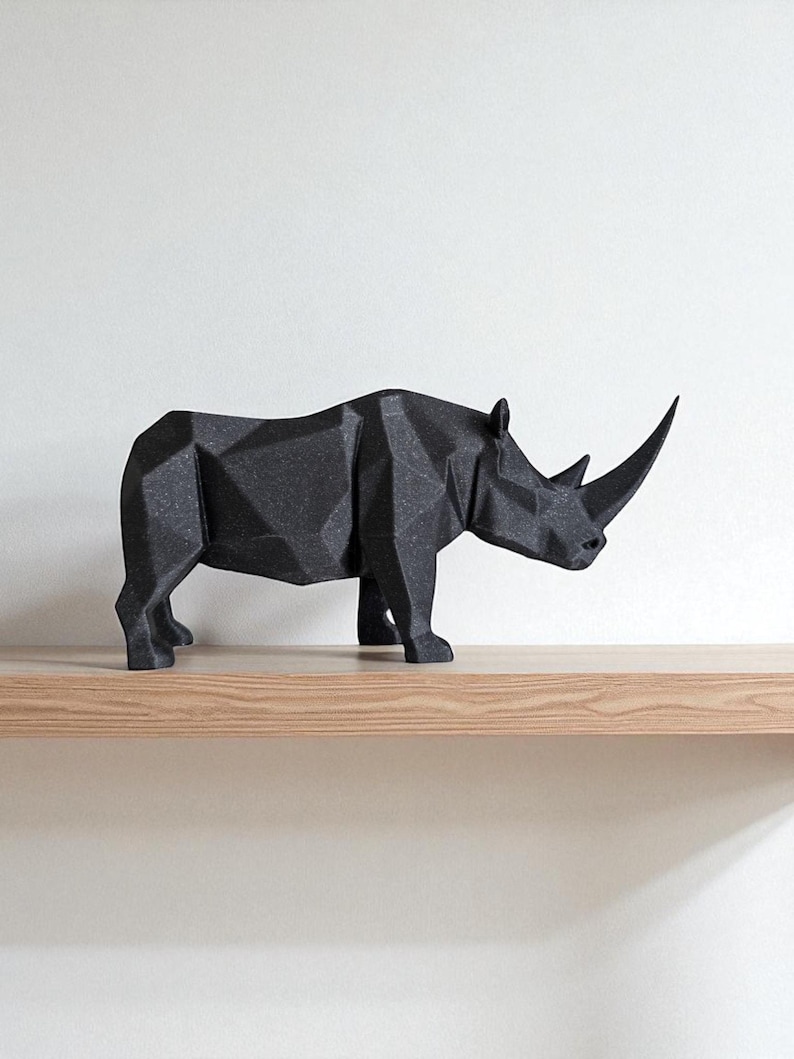 May include: A black, geometric rhino sculpture with a textured surface. The rhino is positioned on a light-colored wooden shelf against a white wall. The sculpture has a prominent horn and is a modern decorative piece.