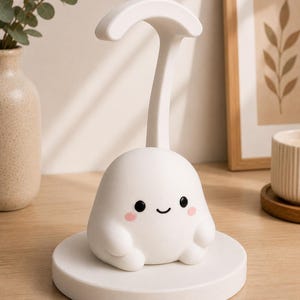 May include: A white, cartoon-like night light with a smiling face and pink cheeks. The light has a curved handle and sits on a round base. The lamp is on a wooden surface.