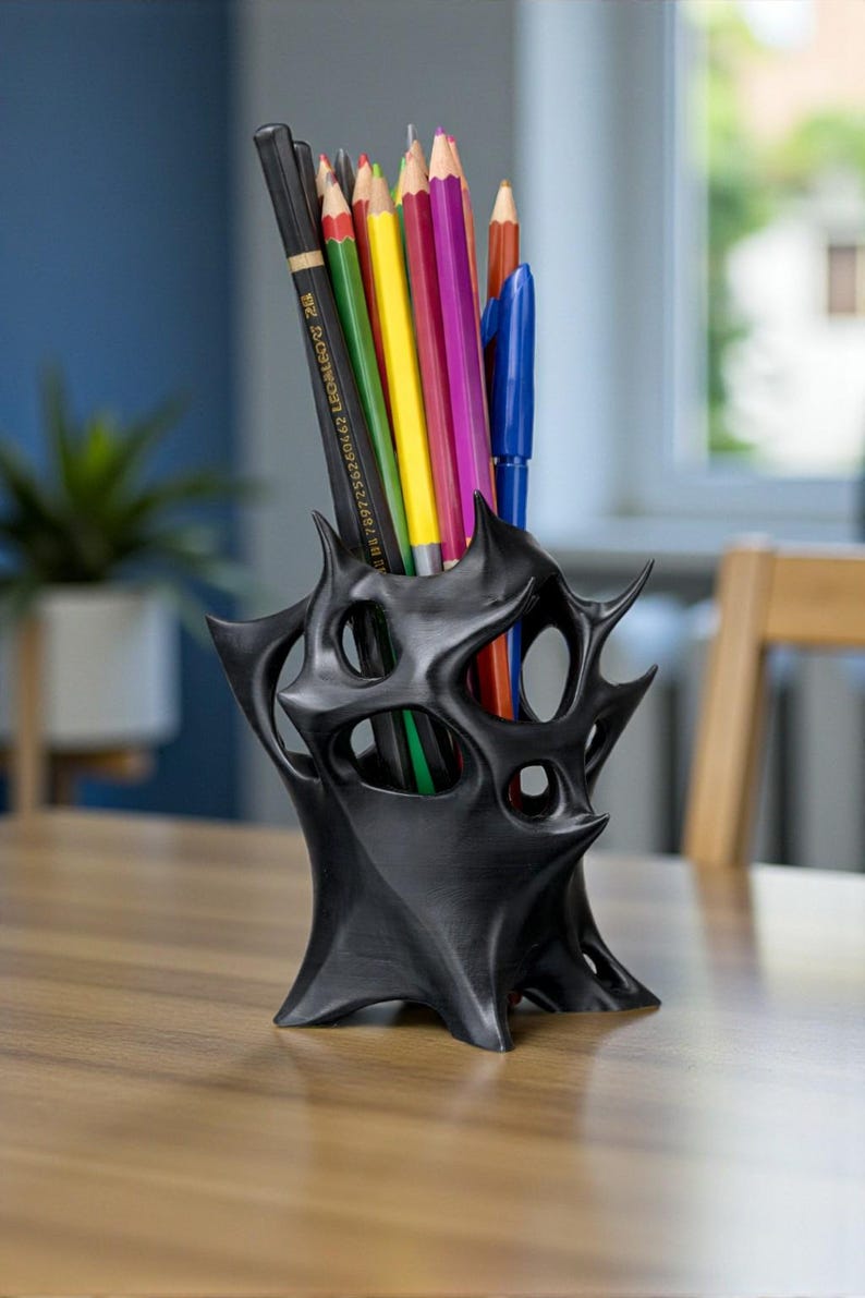 May include: A black, abstract-shaped desk organizer holding a variety of colored pencils and a blue pen. The organizer has a unique, spiky design with multiple openings. The pencils are various colors, including yellow, green, red, and purple.