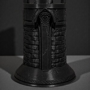 May include: A black, 3D-printed castle tower with a detailed, textured surface. The tower features a crenelated top, arched doorway, and brick-like pattern. The base is round and has a decorative, ridged edge. The tower is approximately 4 inches tall.