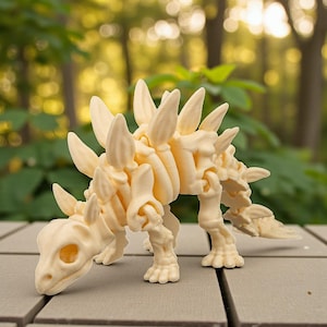 May include: A cream-colored Stegosaurus dinosaur skeleton model. The model features detailed bone structures, including the distinctive plates along its back and a small head. The dinosaur is posed on a gray surface with a blurred green background.
