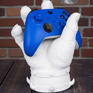 May include: A white, sculpted hand-shaped gaming controller holder. The holder cradles a blue gaming controller. The holder has a textured surface and is set against a brick wall and a dark wooden table.