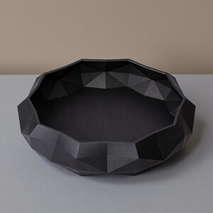 K&ouml;nnte beinhalten: Eine matte, schwarze Dekoschale mit geometrischem, facettiertem Design. Die Schale hat eine breite, flache Form mit glattem Innenraum und strukturierter Au&szlig;enseite, geeignet f&uuml;r kleine Gegenst&auml;nde oder als dekoratives Element.