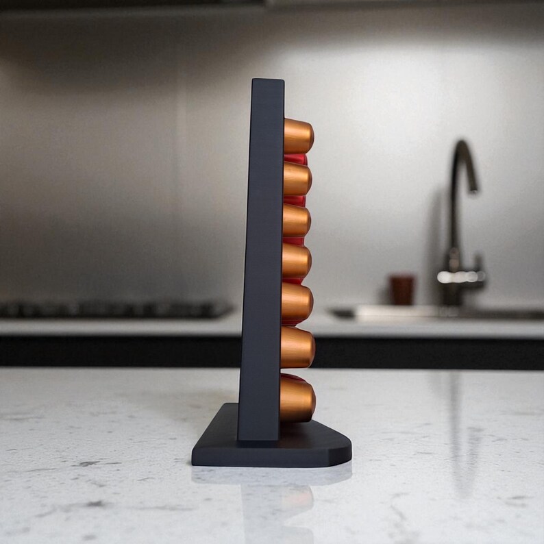 May include: A dark gray coffee pod holder with a rectangular design, holding several copper-colored coffee pods. The holder is displayed on a white countertop, showcasing a modern kitchen aesthetic.