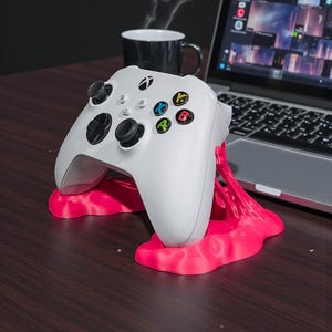 May include: A white Xbox controller rests on a bright pink stand. The controller has black joysticks and buttons with colored accents. A laptop and a black mug are in the background, suggesting a gaming setup.