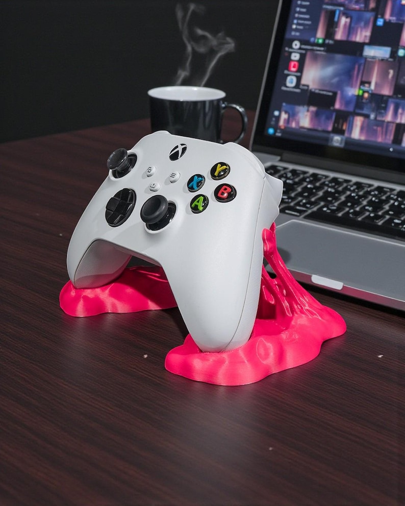 May include: A white Xbox controller rests on a bright pink stand. The controller has black joysticks and buttons with colored accents. A laptop and a black mug are in the background, suggesting a gaming setup.