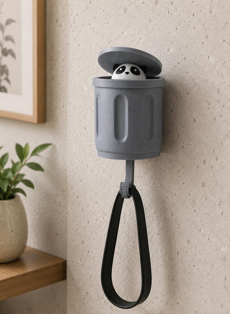 May include: A gray panda-themed wall-mounted trash can with a black handle. The panda's head peeks out from the open lid. The trash can is made of plastic and is designed to hold small items. The handle is attached to the bottom of the can.