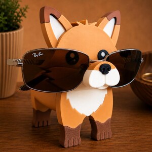 May include: A whimsical orange, brown, and white fox-shaped eyeglass holder. The fox is wearing a pair of Ray-Ban sunglasses with a silver frame and dark lenses. The holder is made of wood and sits on a wooden surface.