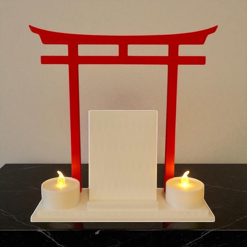 May include: A red Torii gate-shaped candle holder with a white base. Two battery-operated tea light candles sit on the base. A white rectangular panel is centered within the gate. The background is a neutral colour.