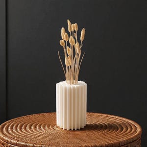 May include: A white, ribbed ceramic vase holding dried wheat stalks. The vase is cylindrical and sits on a woven, brown surface. The wheat stalks are a light tan color, contrasting with the white vase.