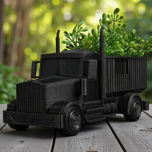 Puede incluir: Una jardinera decorativa negra con forma de camión, con una caja de carga llena de follaje verde. El camión presenta ruedas detalladas, una rejilla delantera y tubos de escape. La jardinera está hecha de un material sólido y se encuentra sobre una superficie de madera. Aproximadamente 30 cm de largo.